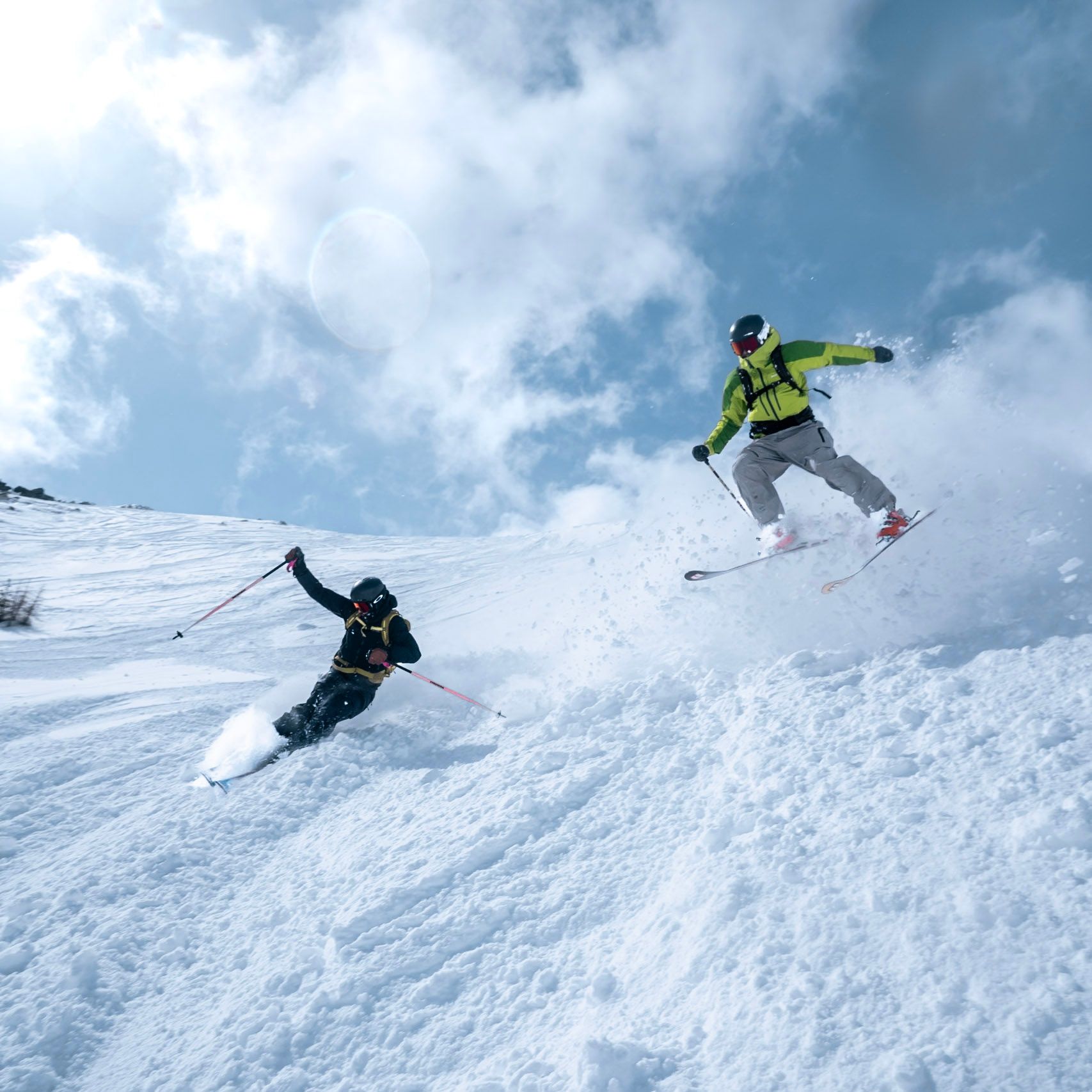 Zwei Skifahrer im Tiefschnee, einer davon in der Luft vor bewölktem Himmel